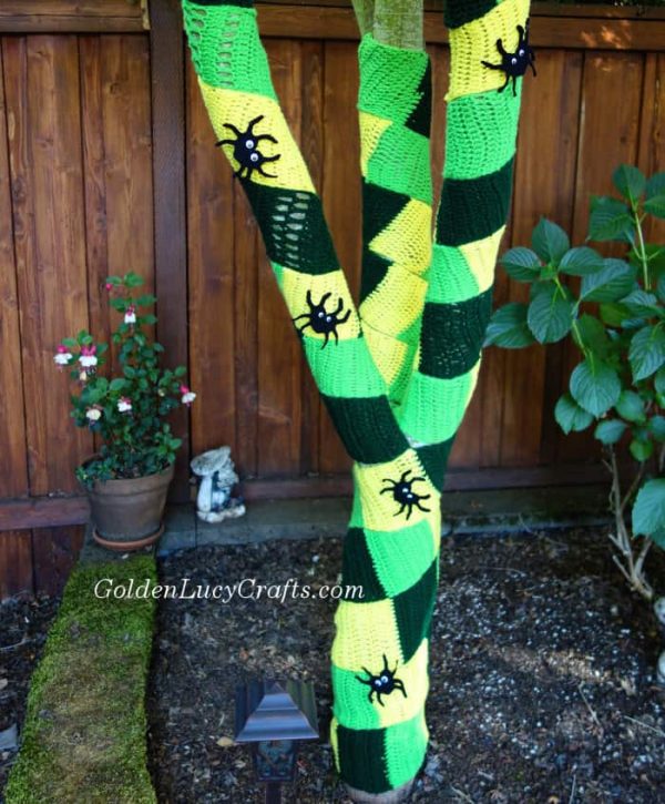 Tree wrapped in green and yellow crocheted pieces, embellished with small crocheted spiders.
