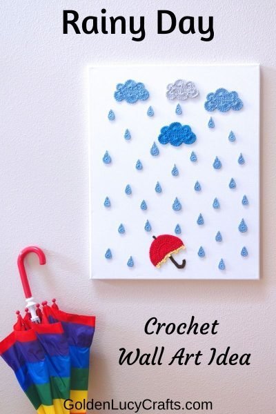 Rainy Day crochet wall art hanging on the wall - clouds, raindrops, umbrella appliques on canvas, umbrella in rainbow colors next to it.