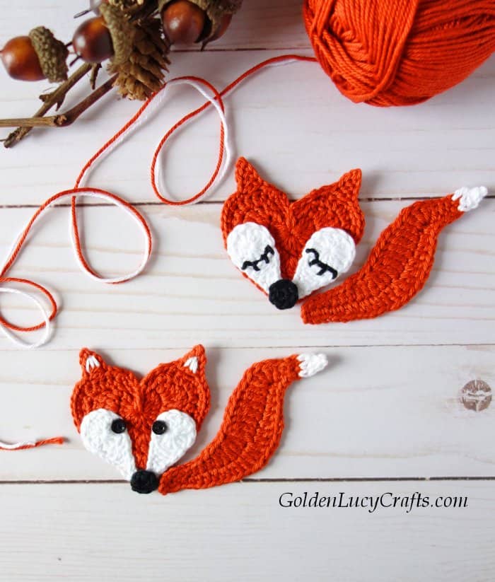 Two crocheted fox appliques, ball of orange yarn, acorns in the background.