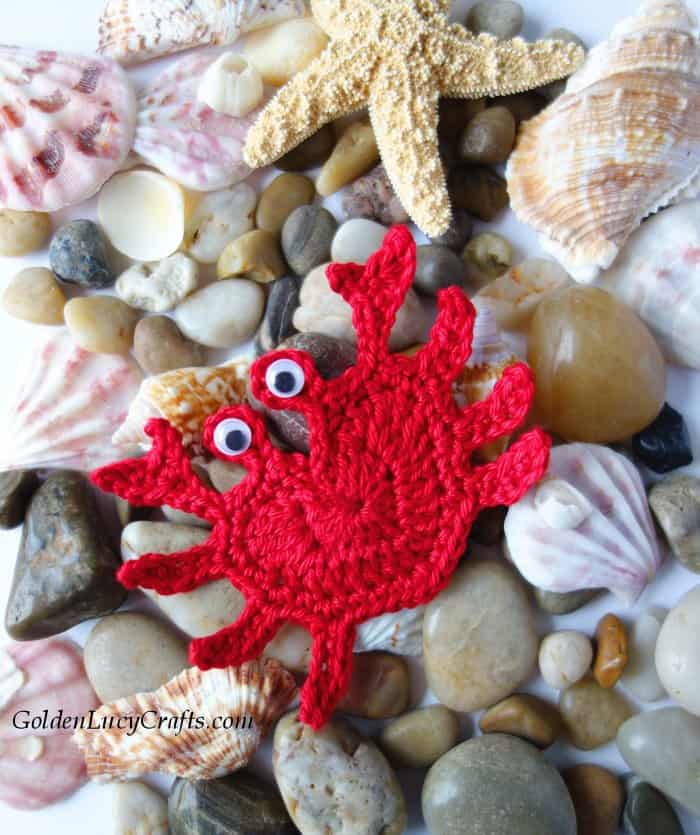 Crochet red crab applique laying on top of the rocks and sea shells.
