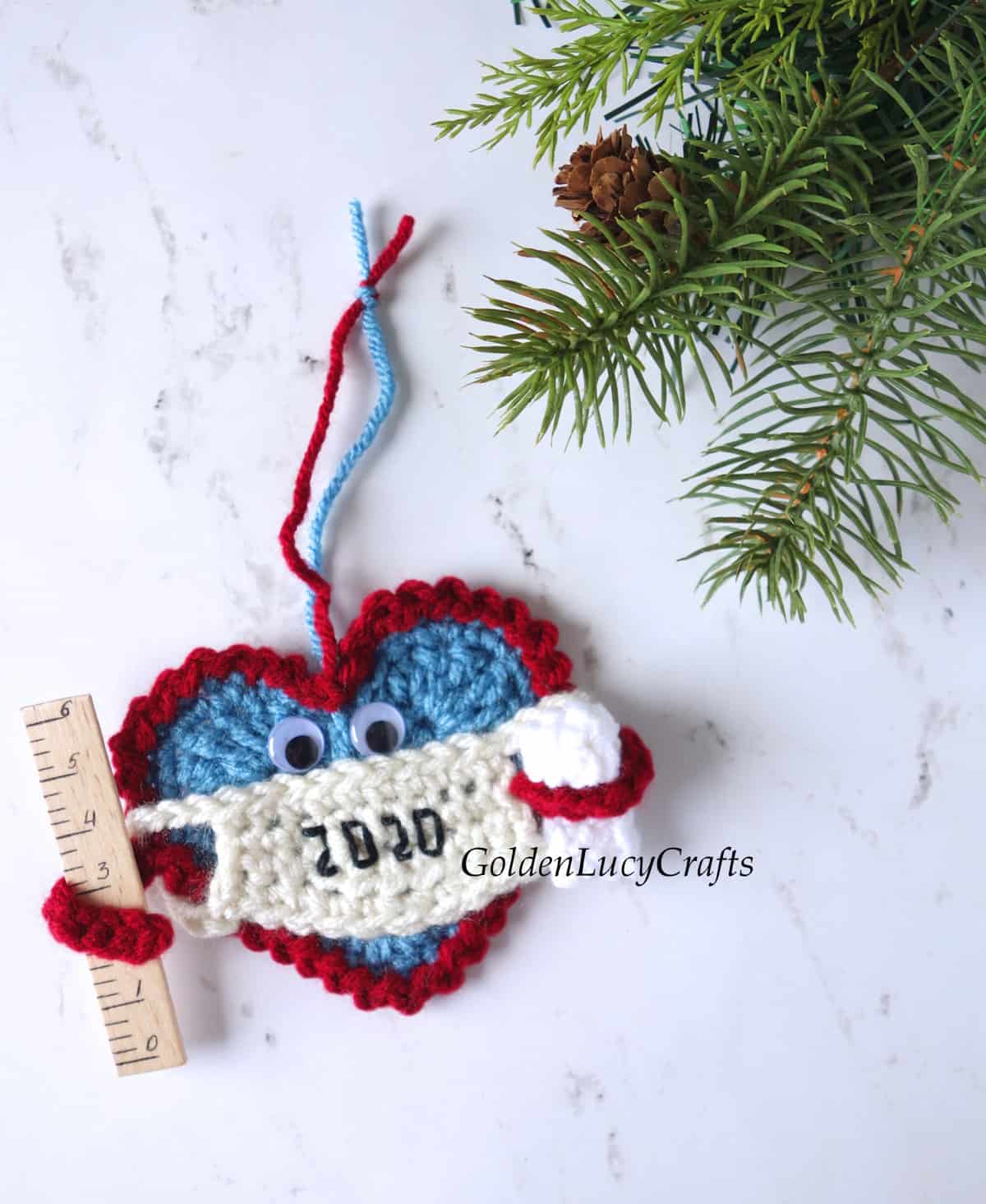 Crochet Christmas 2020 ornament. The ornament is a Heart wearing a mask and holding a ruler in one arm and a toilet paper roll in the other.