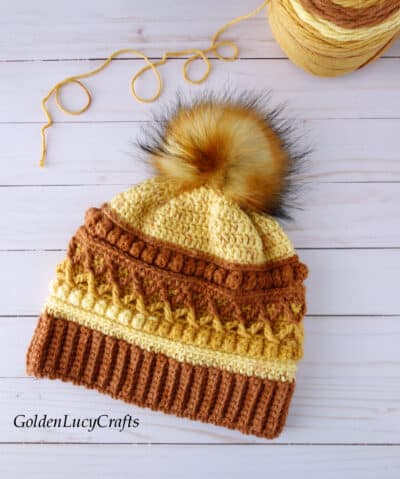 Crocheted hat in brown, orange and yellow colors embellished with faux fur pompom, ball of yarn in the background.
