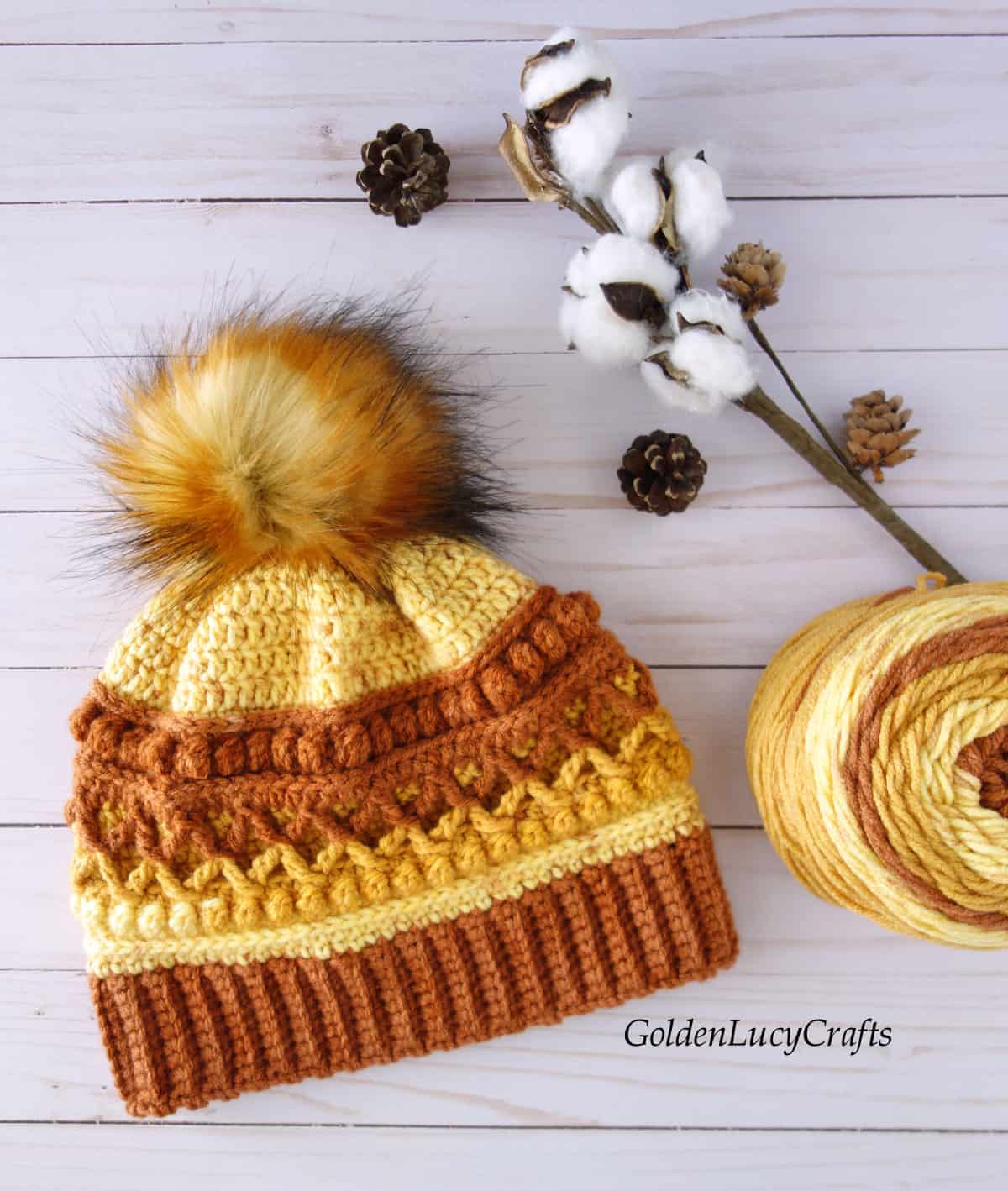 Crocheted hat in brown, orange and yellow colors embellished with fur pompom, ball of yarn, cotton branch, pine cones are laying on the flat surface.