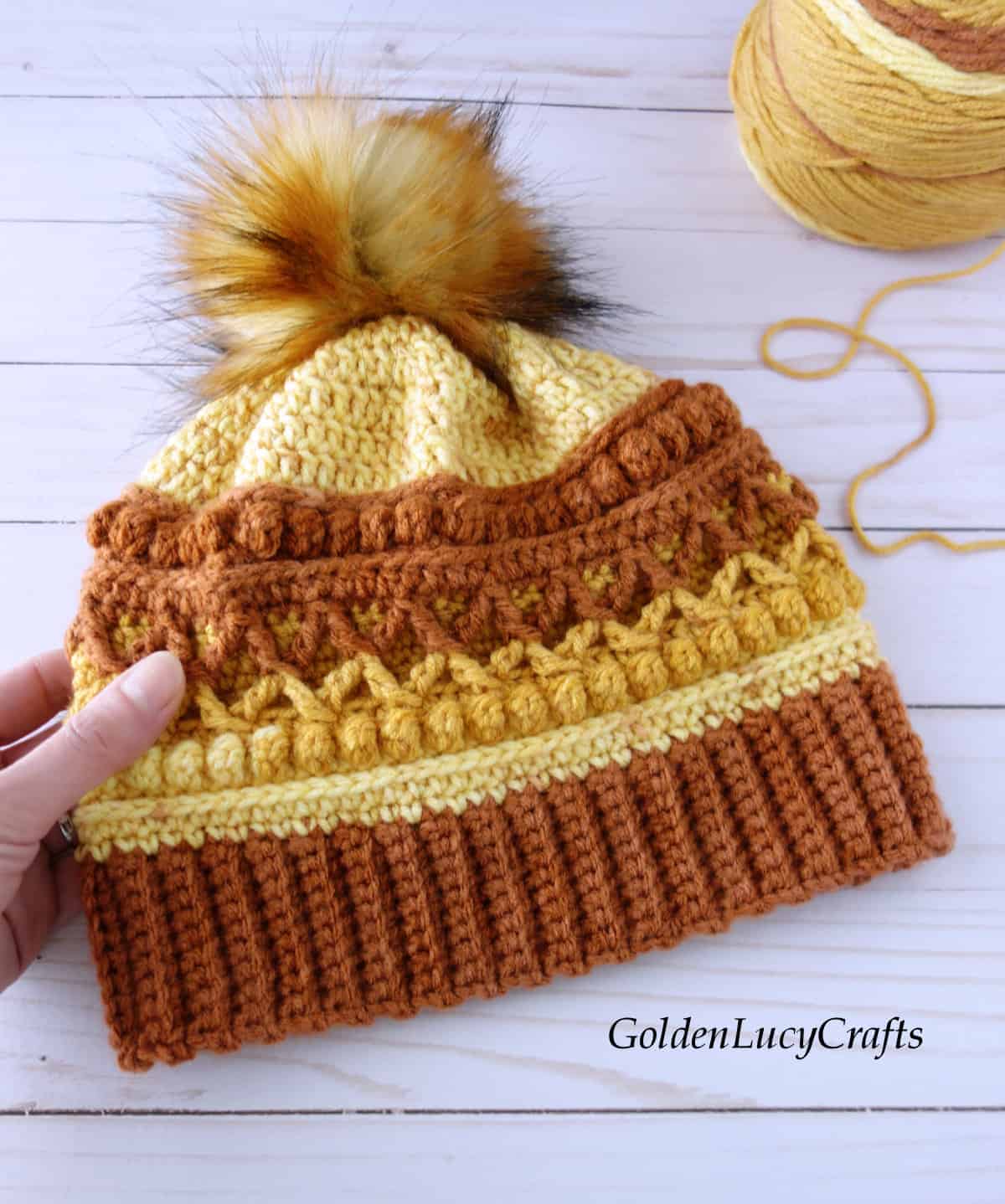 Hand holding crocheted hat in brown, orange and yellow colors embellished with faux fur pompom, ball of yarn in the background.