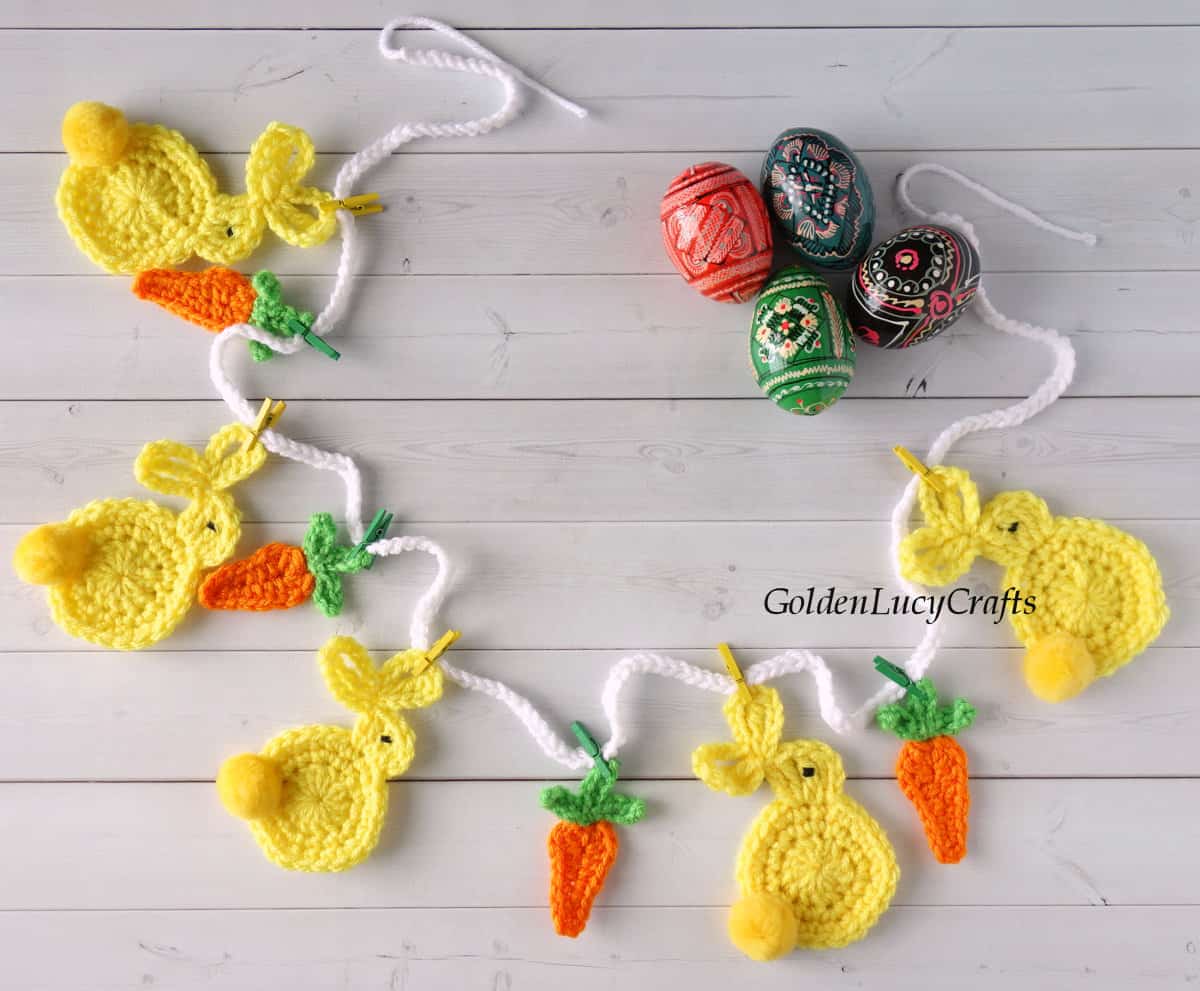 Crocheted Easter garland with bunnies and carrots laying on surface with painted Easter eggs next to it.