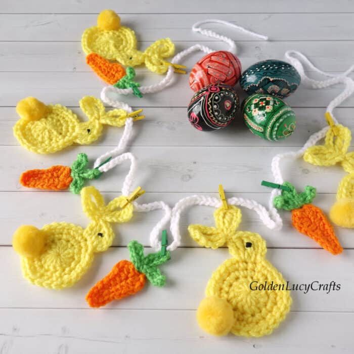 Crocheted Easter garland with bunnies and carrots laying on surface with painted Easter eggs next to it.