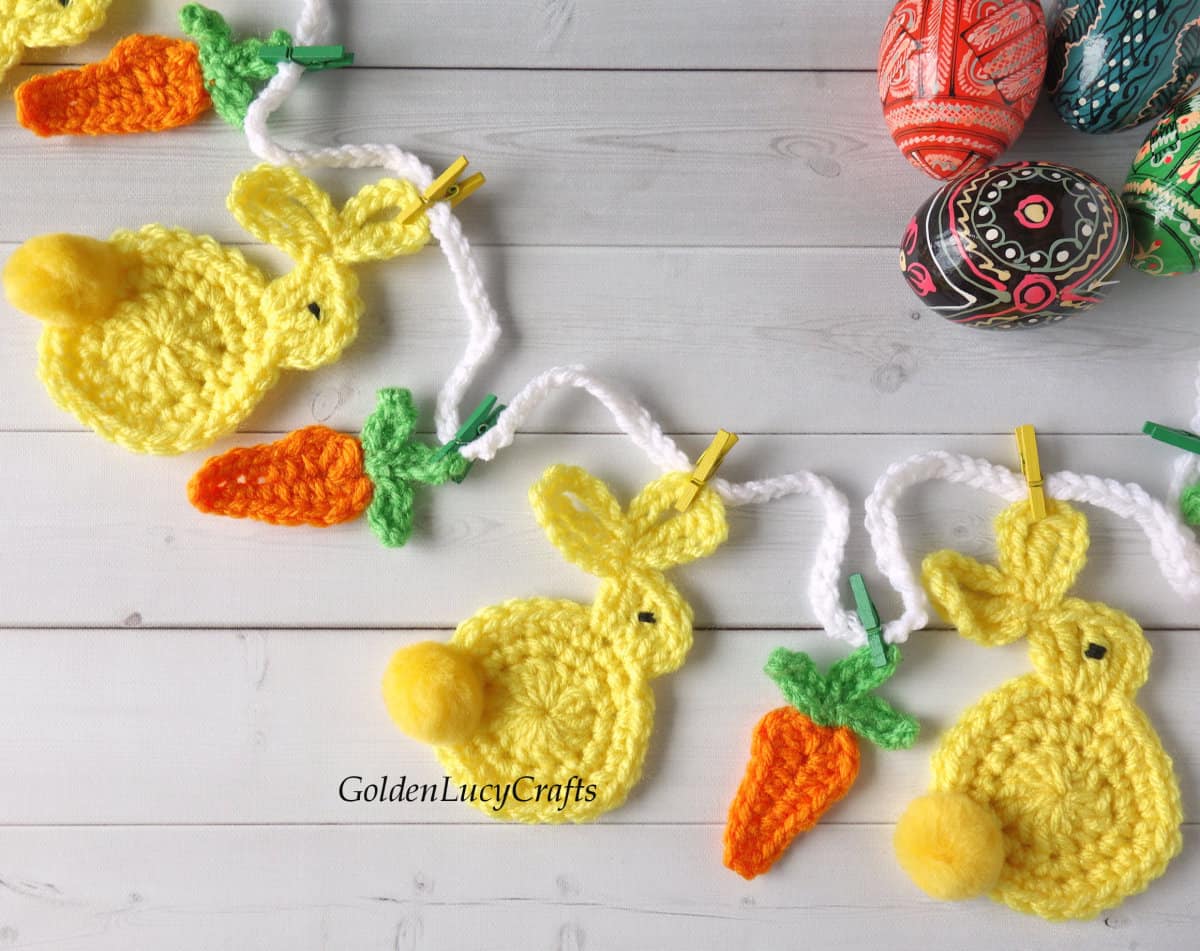 Crocheted Easter garland with bunnies and carrots close up picture, painted Easter eggs in the background.