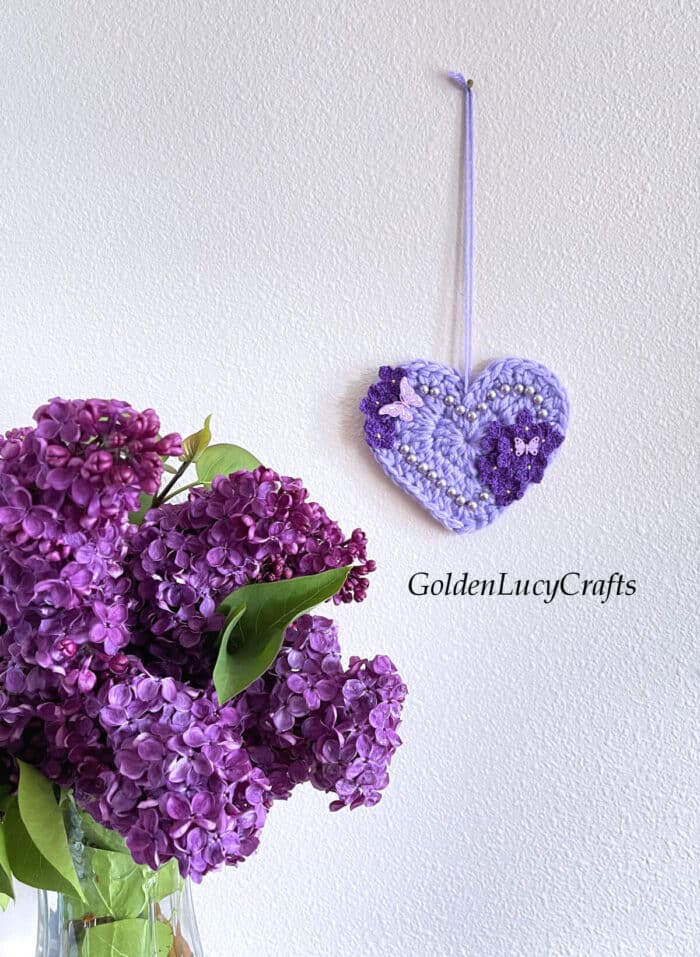 Crocheted heart embellished with crochet lilac flowers and beads hanging on the wall, lilac bouquet in a base next to it.