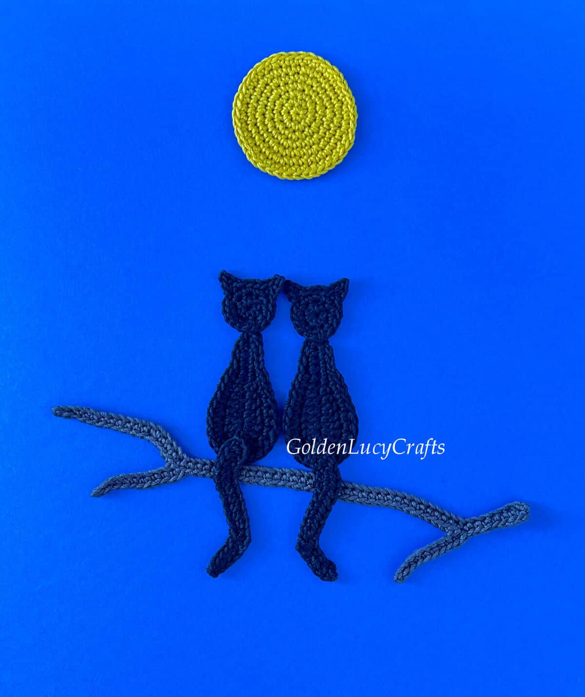Crocheted two black cats sitting on a tree branch under a full moon on dark blue background.