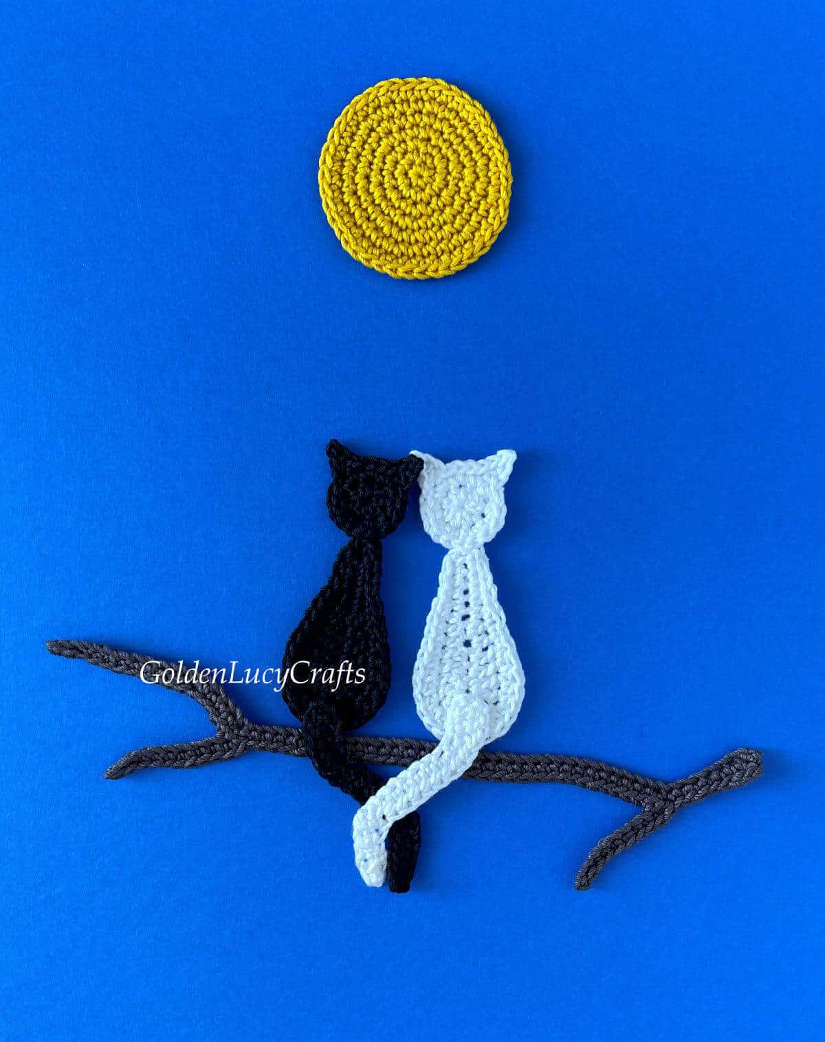 Crocheted two cats in love sitting on a tree branch under a full moon on dark blue background.