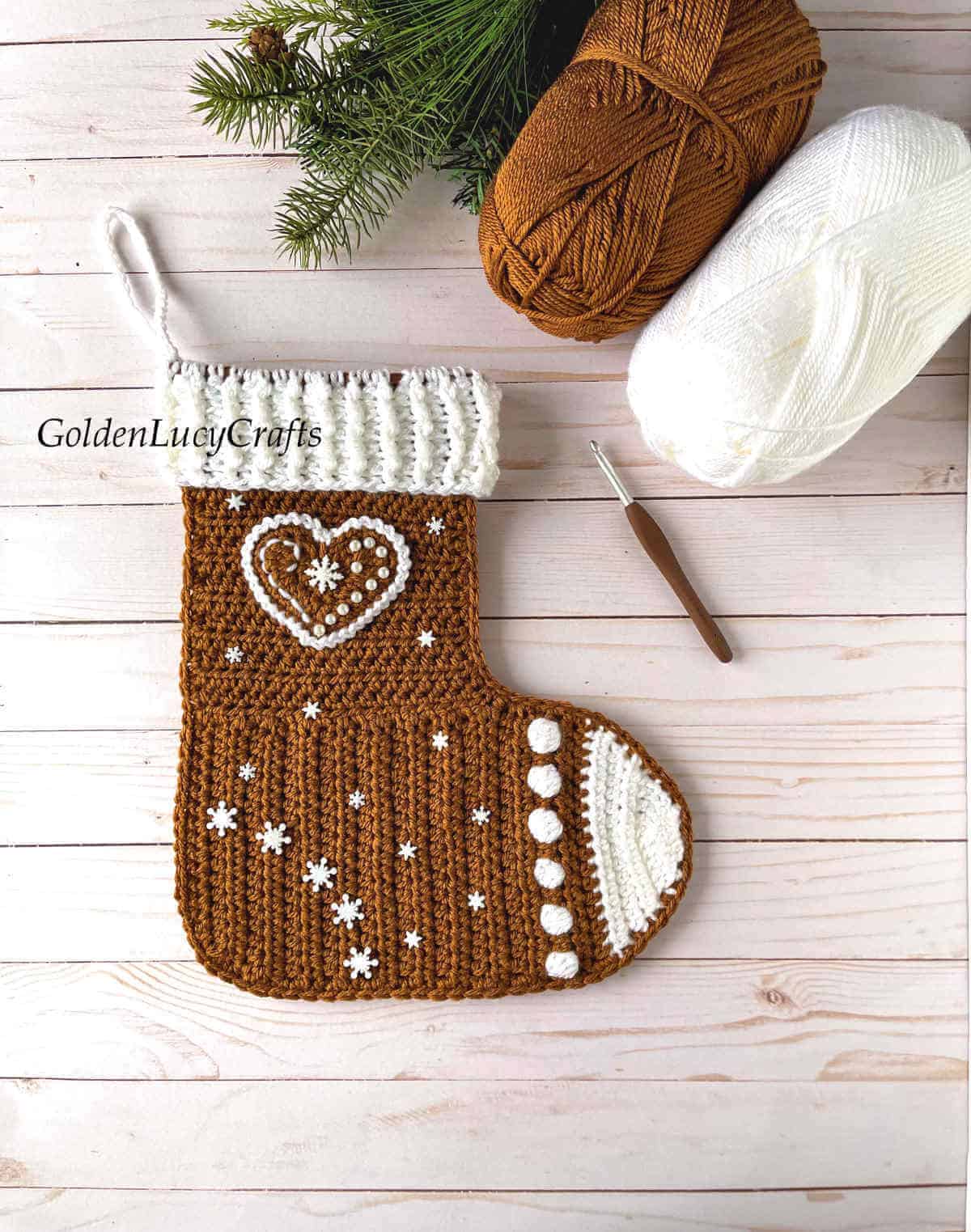 Crochet gingerbread Christmas stocking, crochet hook, two skeins of yarn, Christmas tree branch in the background.