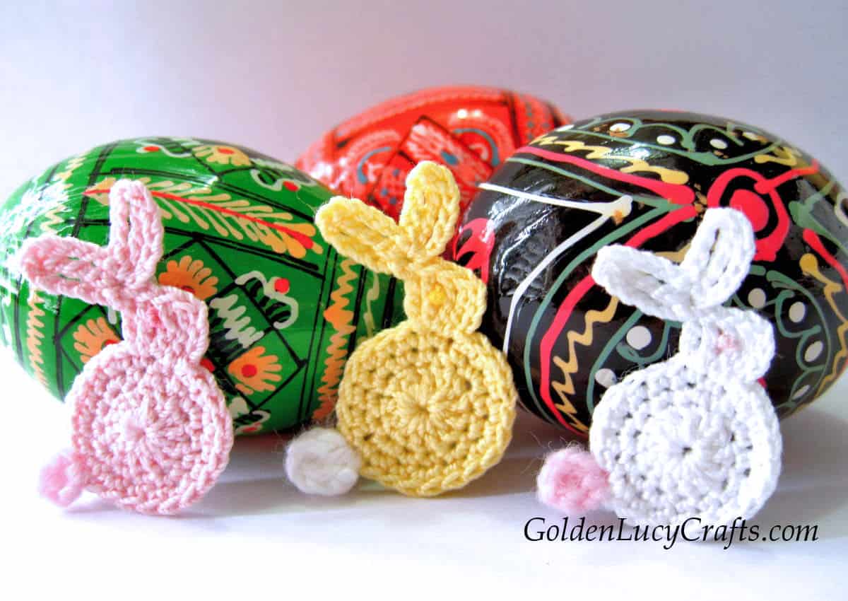 Three crocheted Easter bunny appliques, painted Easter eggs in the background.