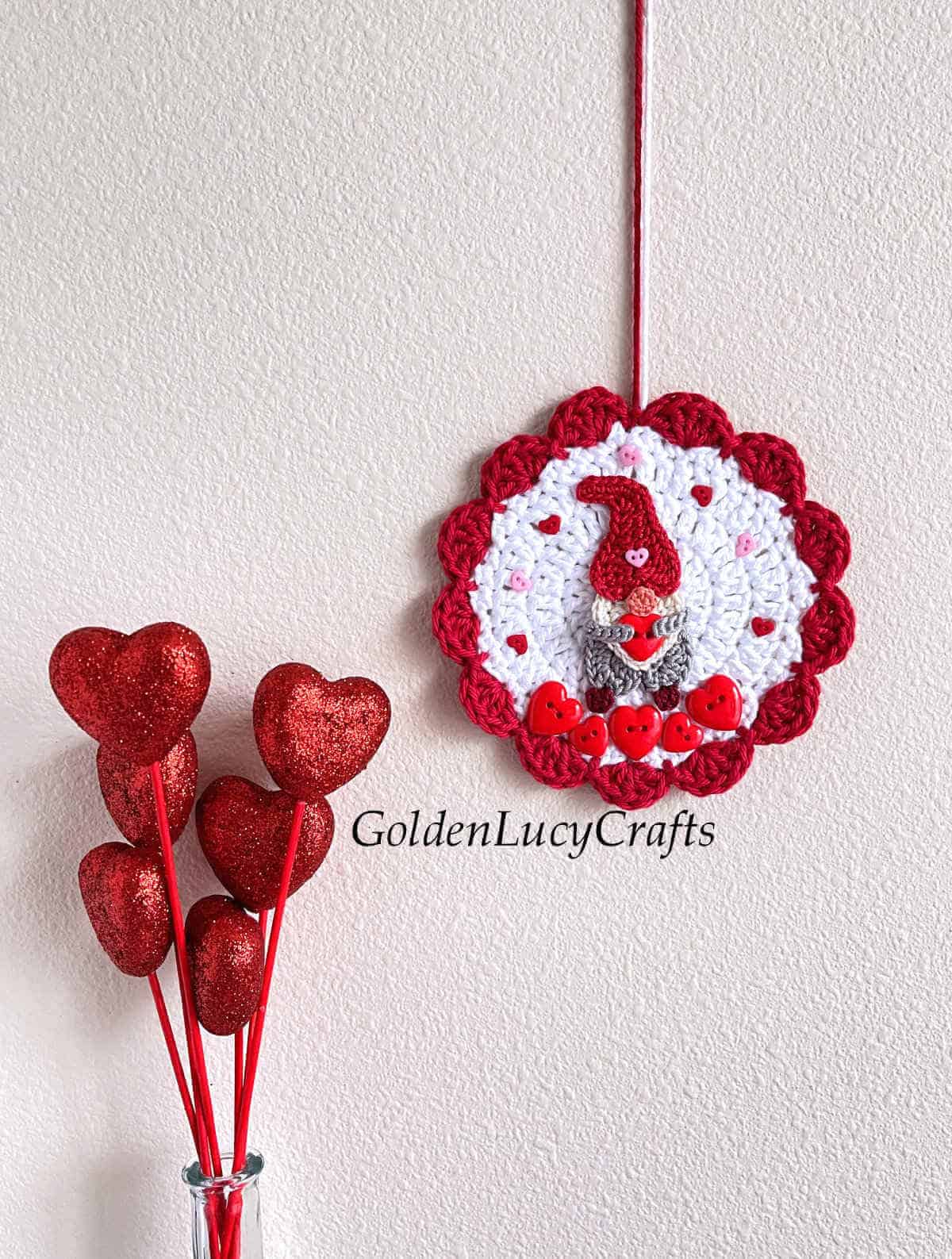 Crochet ornament with gnome on it hanging on the wall, bunch of hearts in a vase next to it.