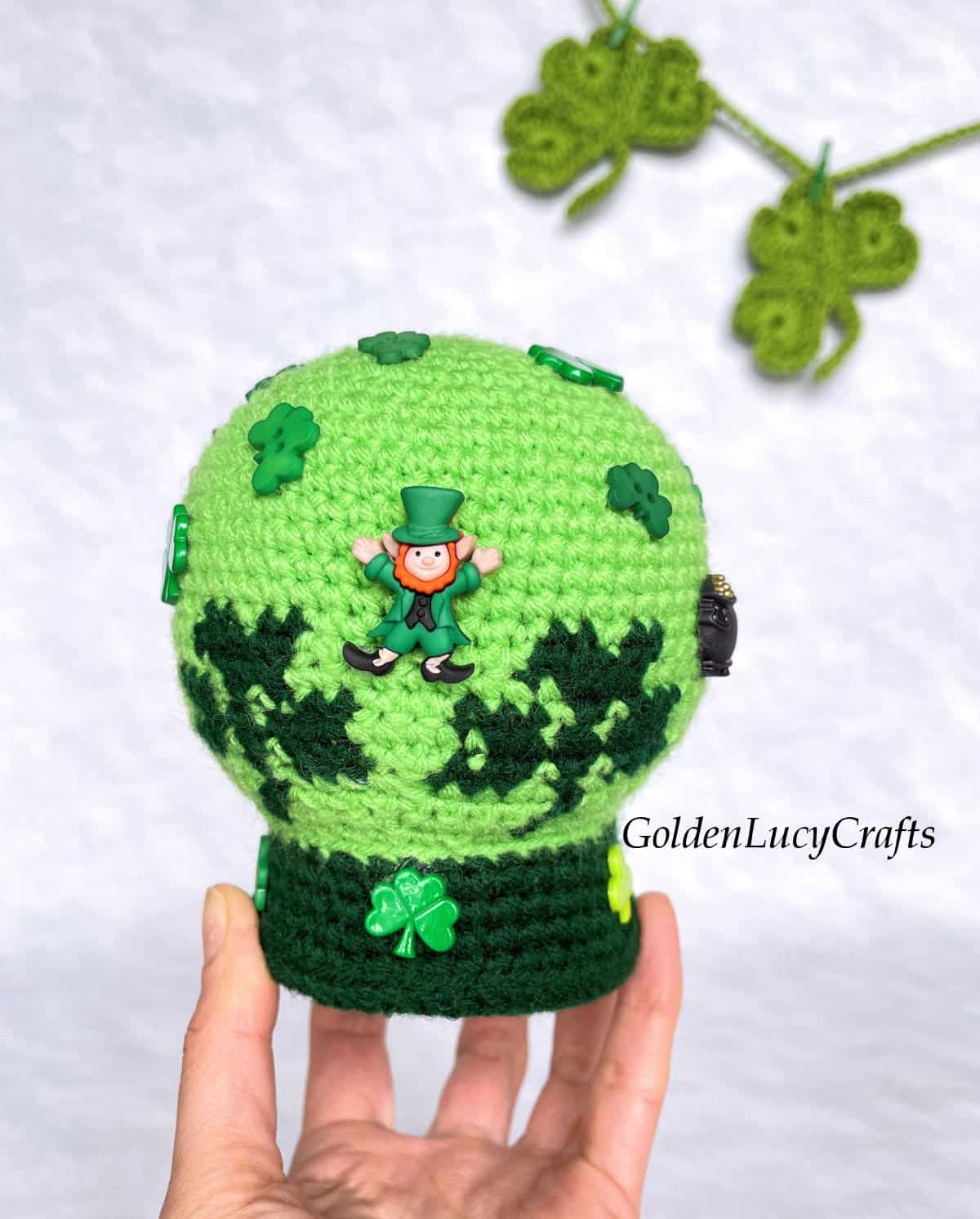 Crochet St. Patrick's Day snow globe amigurumi held by finger tips.