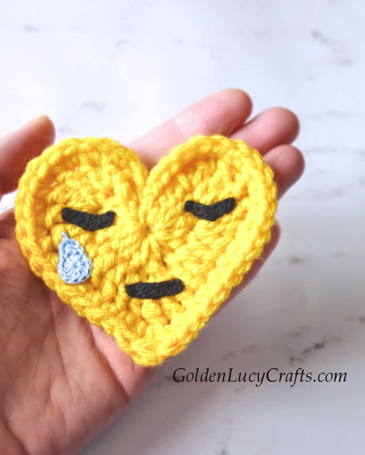 Crocheted crying face emoji applique in the palm of a hand.
