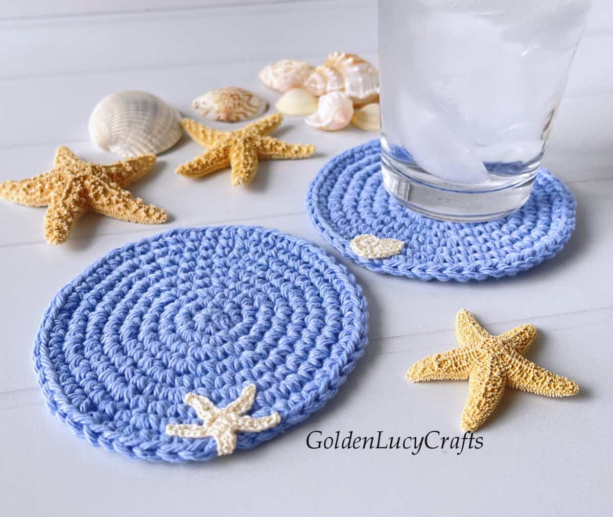 Crocheted ocean themed blue coasters, glass of water, sea stars and sea shells.