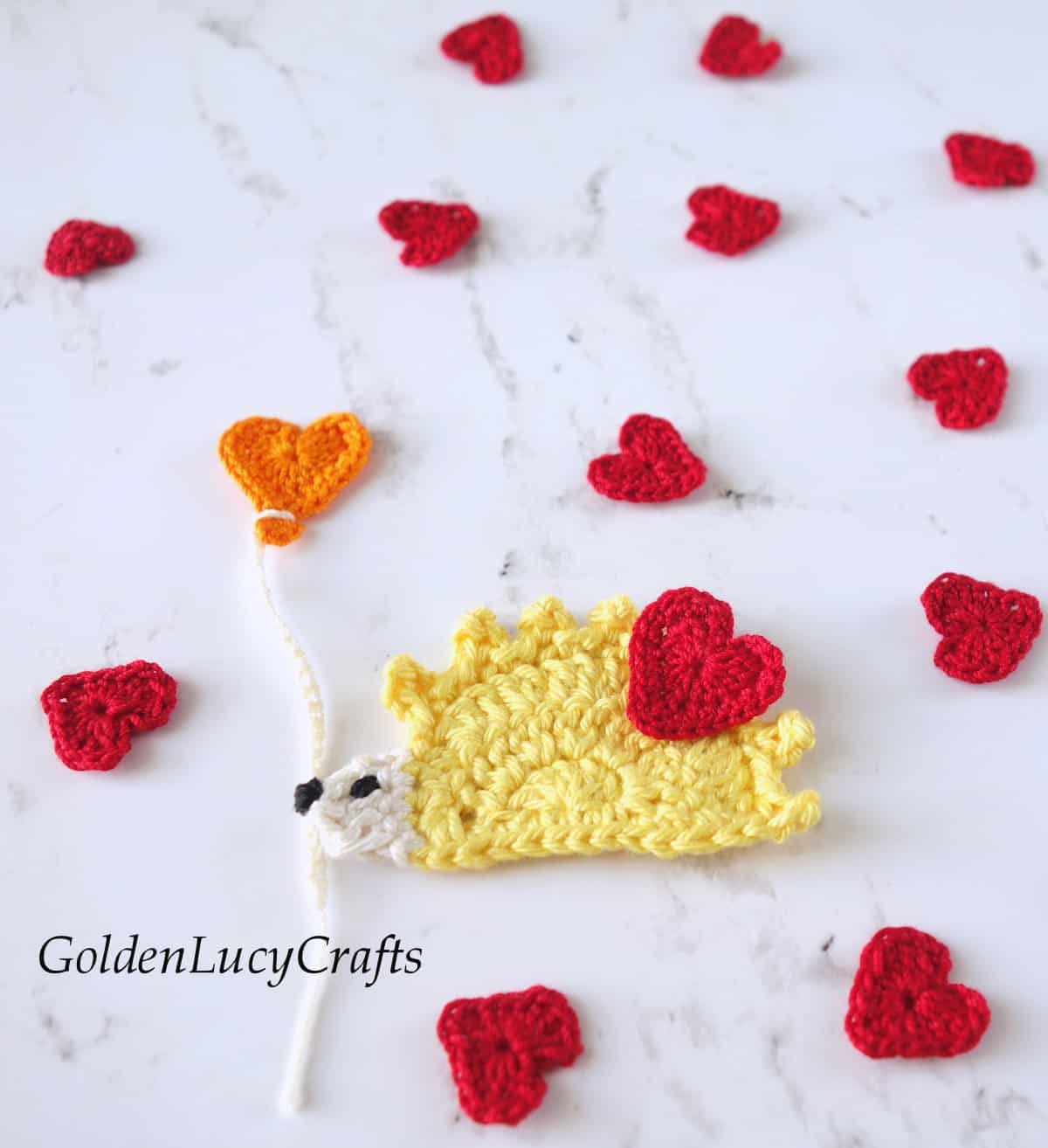 Crochet appliques yellow hedgehog with heart balloon and small red hearts around.