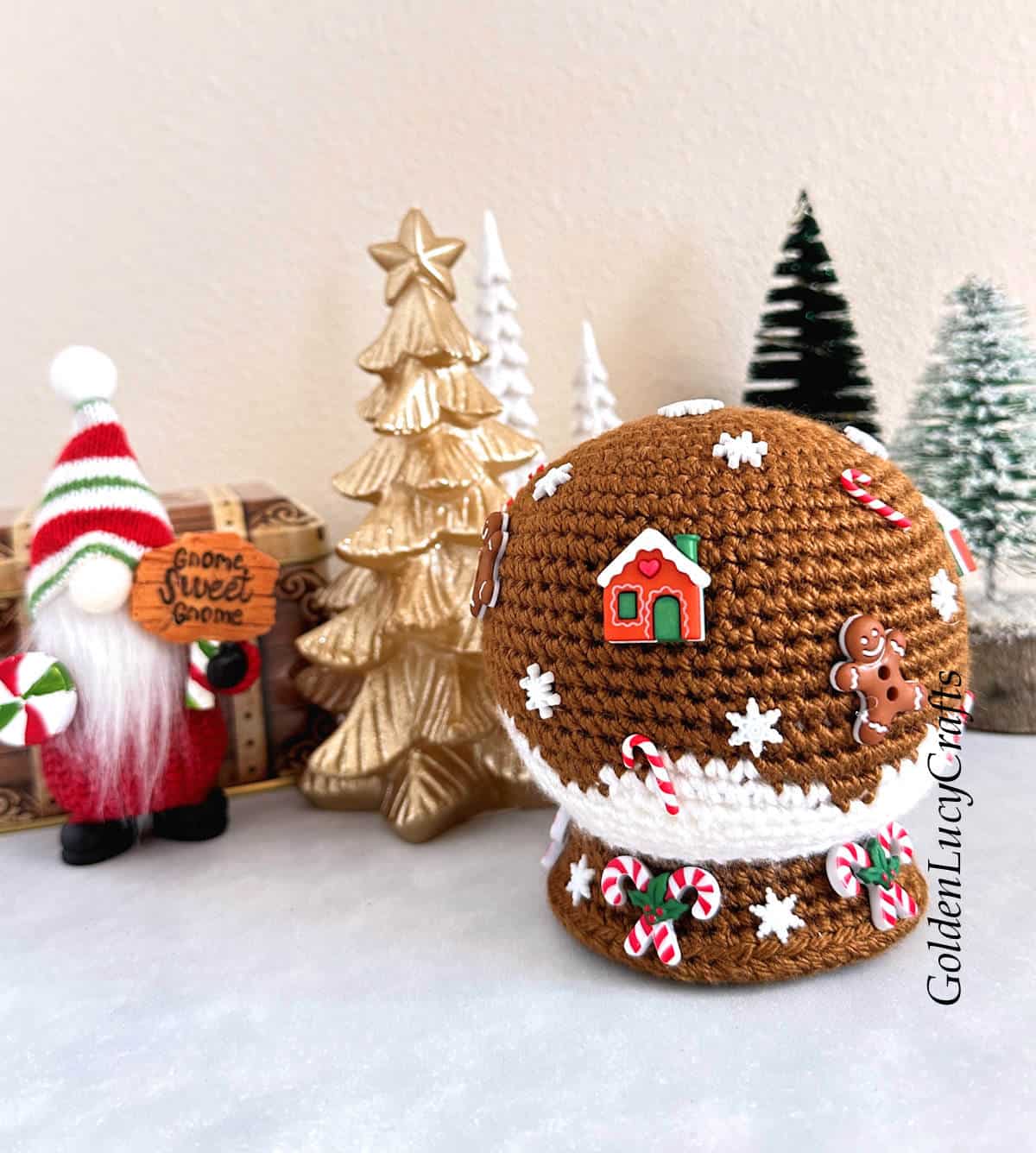 Crochet gingerbread themed snow globe, Christmas trees and gnome in the background.