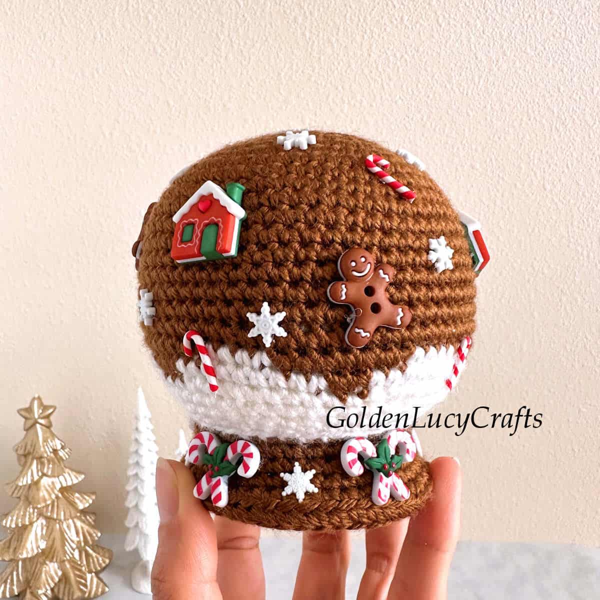 Crocheted gingerbread snow globe amigurumi held by fingertips.