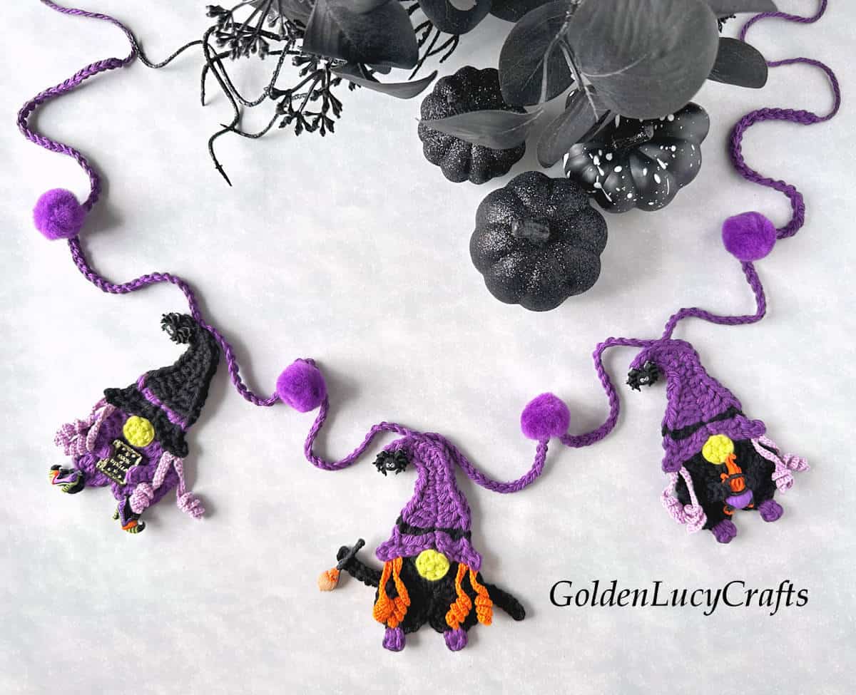 Crochet Halloween garland with witch gnomes and purple pom-poms, black pumpkins and flowers in the background.