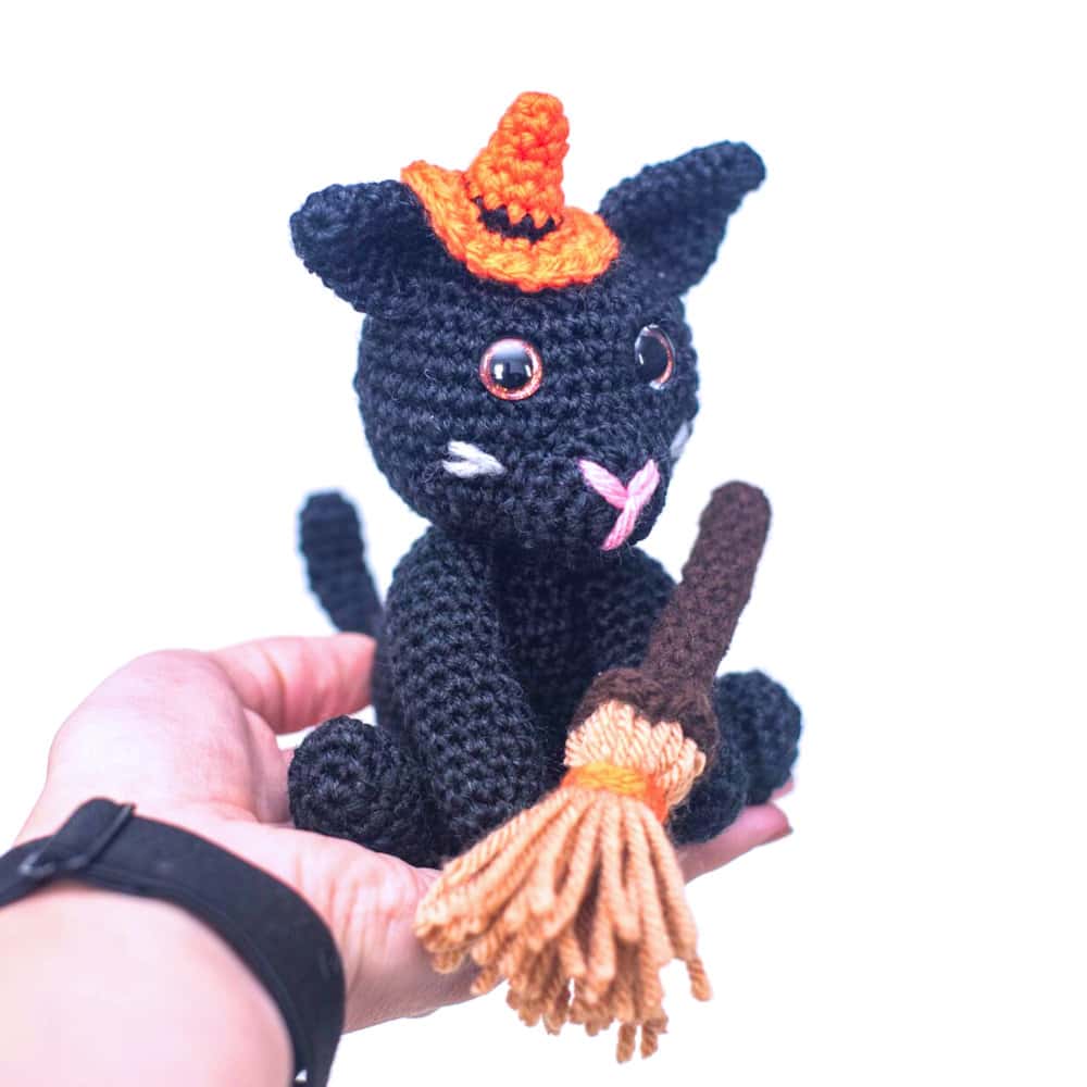 Crocheted cat with broom dressed in orange hat.