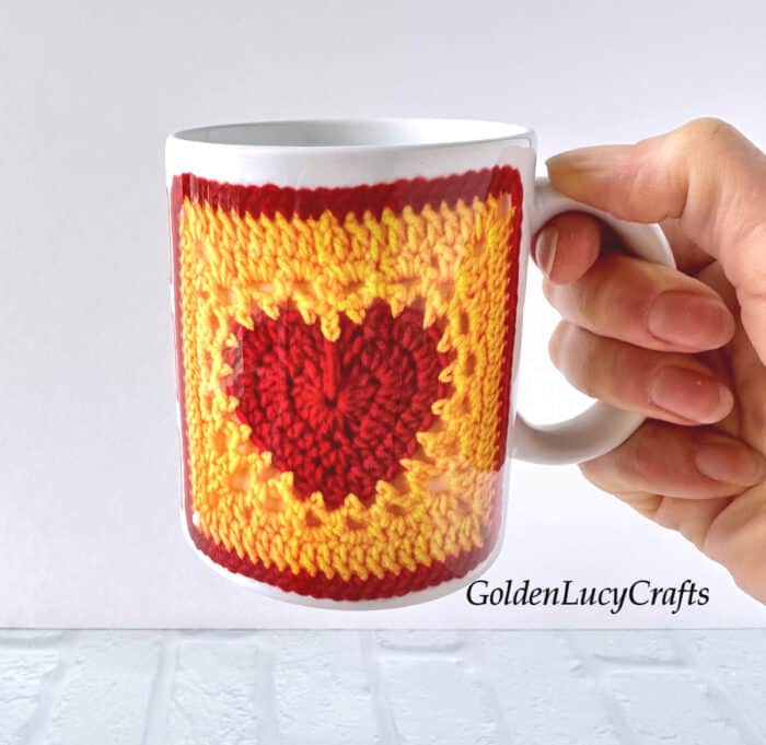 Hand holding a mug with crochet heart square printed on it.