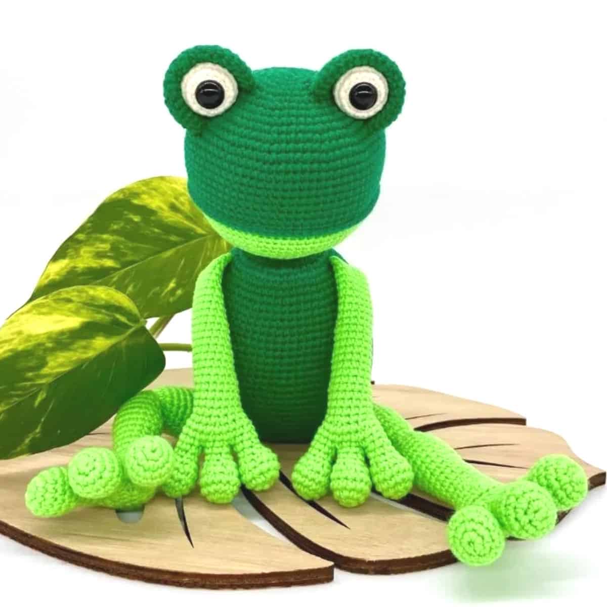 Crocheted frog toy is sitting on a wooden leaf.