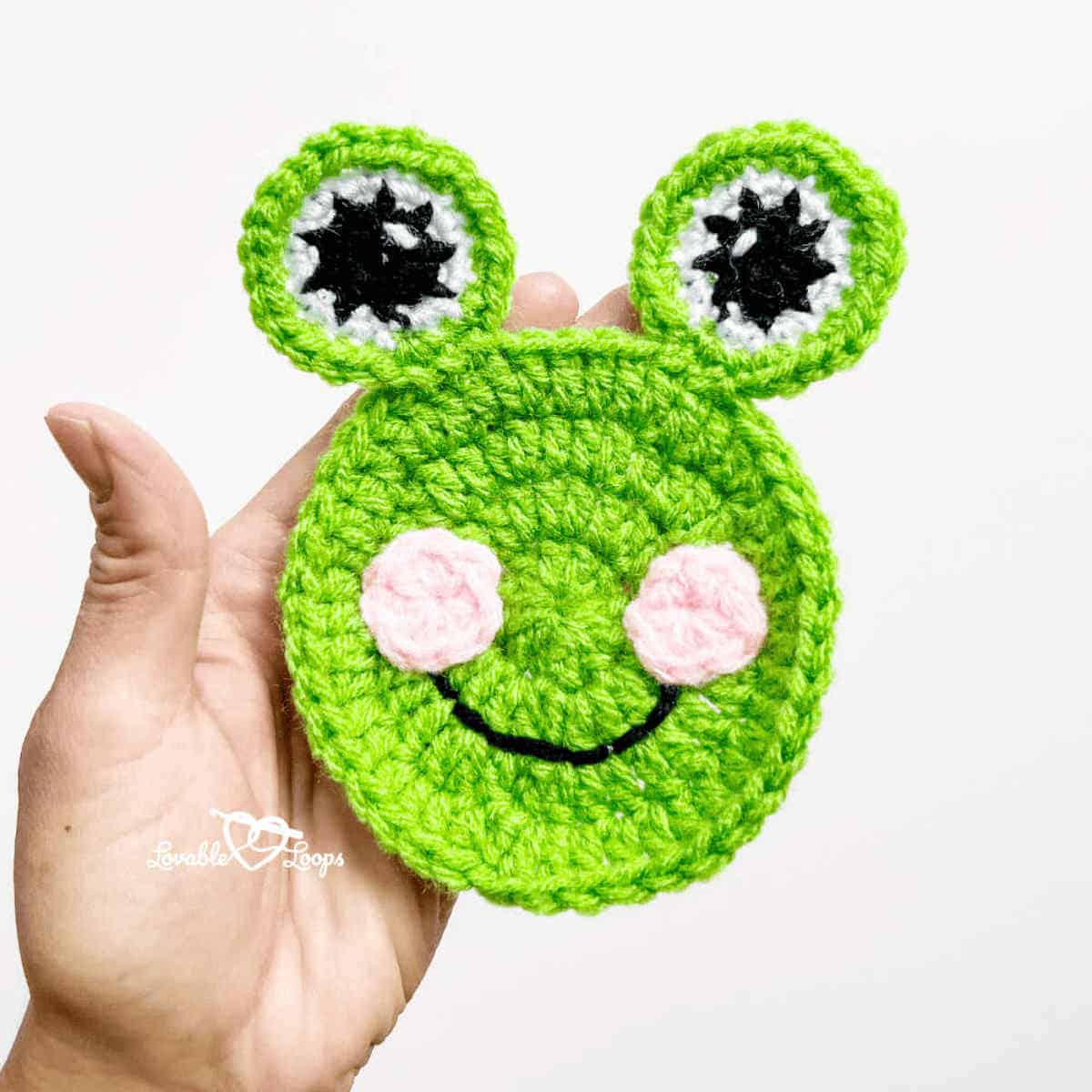 Crocheted frog applique in the palm of a hand.