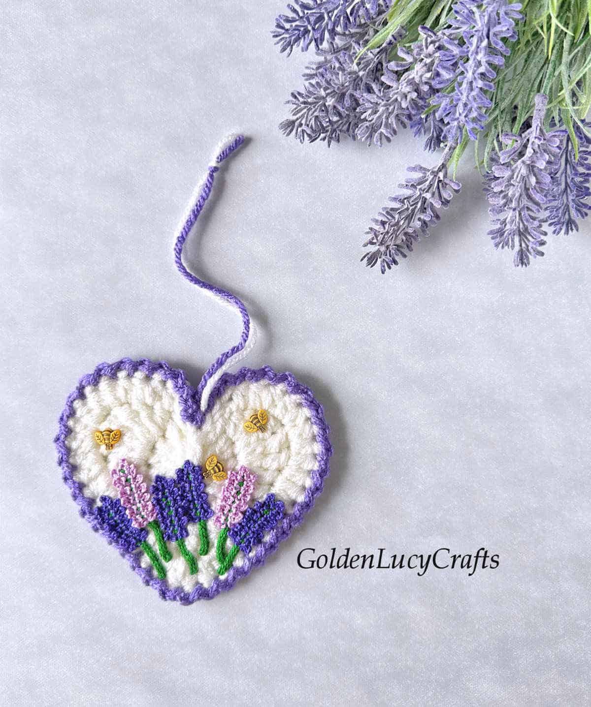 A crocheted heart with lavender flower appliques and bee buttons attached to it.