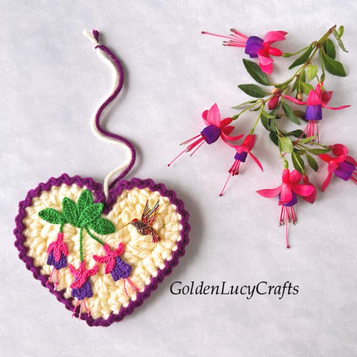 Crocheted heart ornament with fuchsia applique and hummingbird pin on it, real fuchsia flowers in the background.