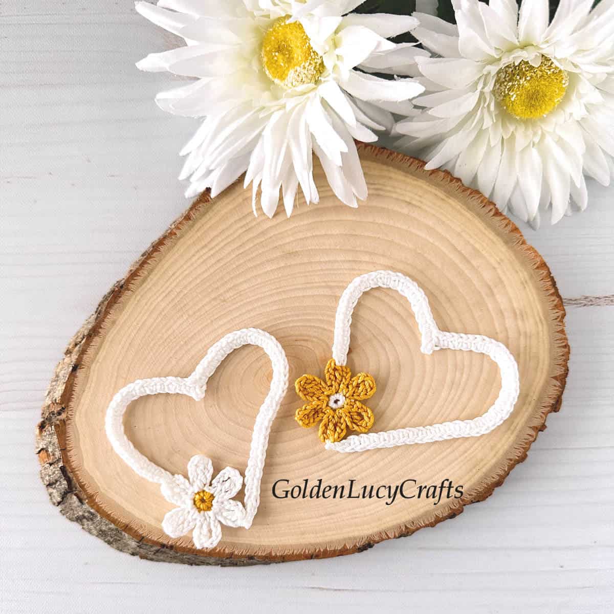 Two crochet white open hearts on wood slice, daisy flowers in the background.