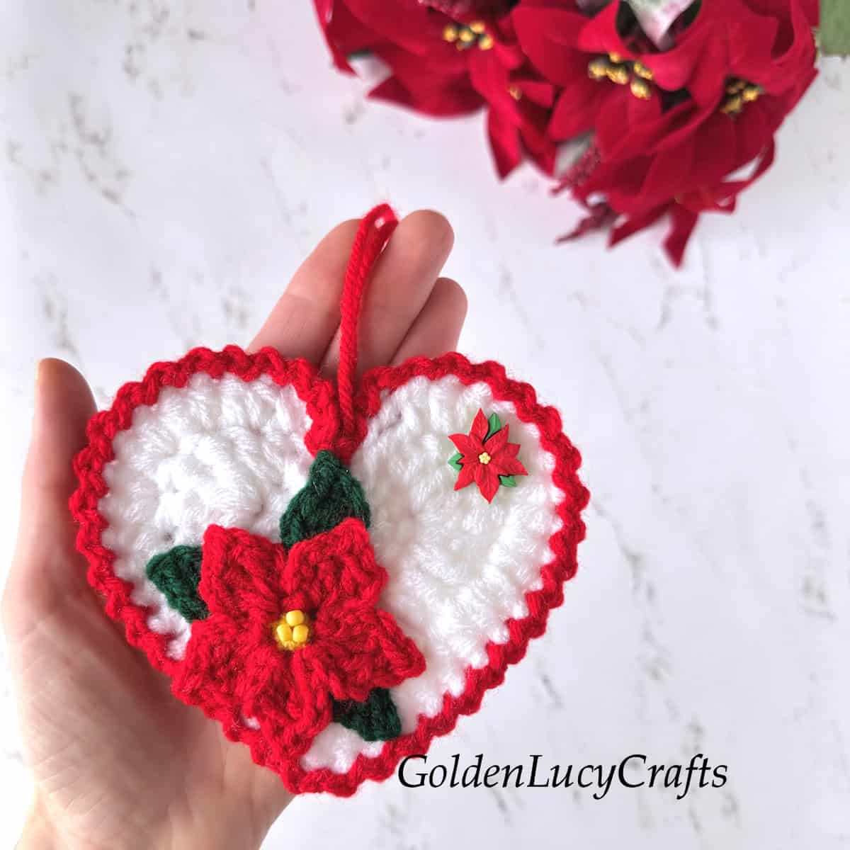 Hand holding a crocheted white heart with a red poinsettia flower on it.