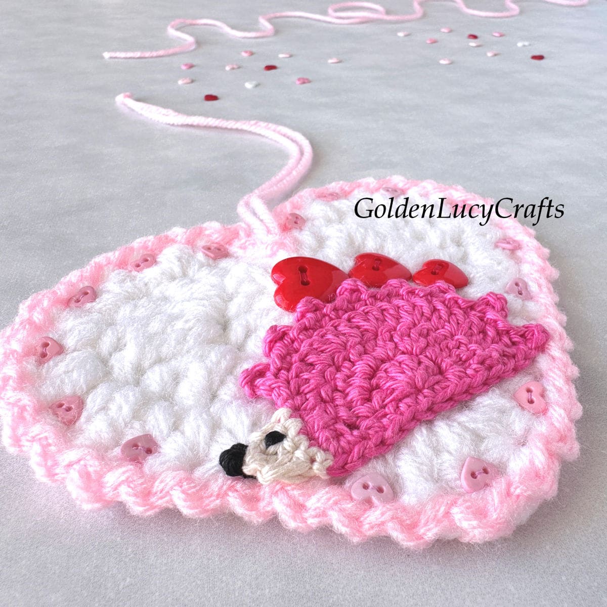 Close up picture of crocheted heart-shaped ornament with pink hedgehog carrying three red hearts on its back.