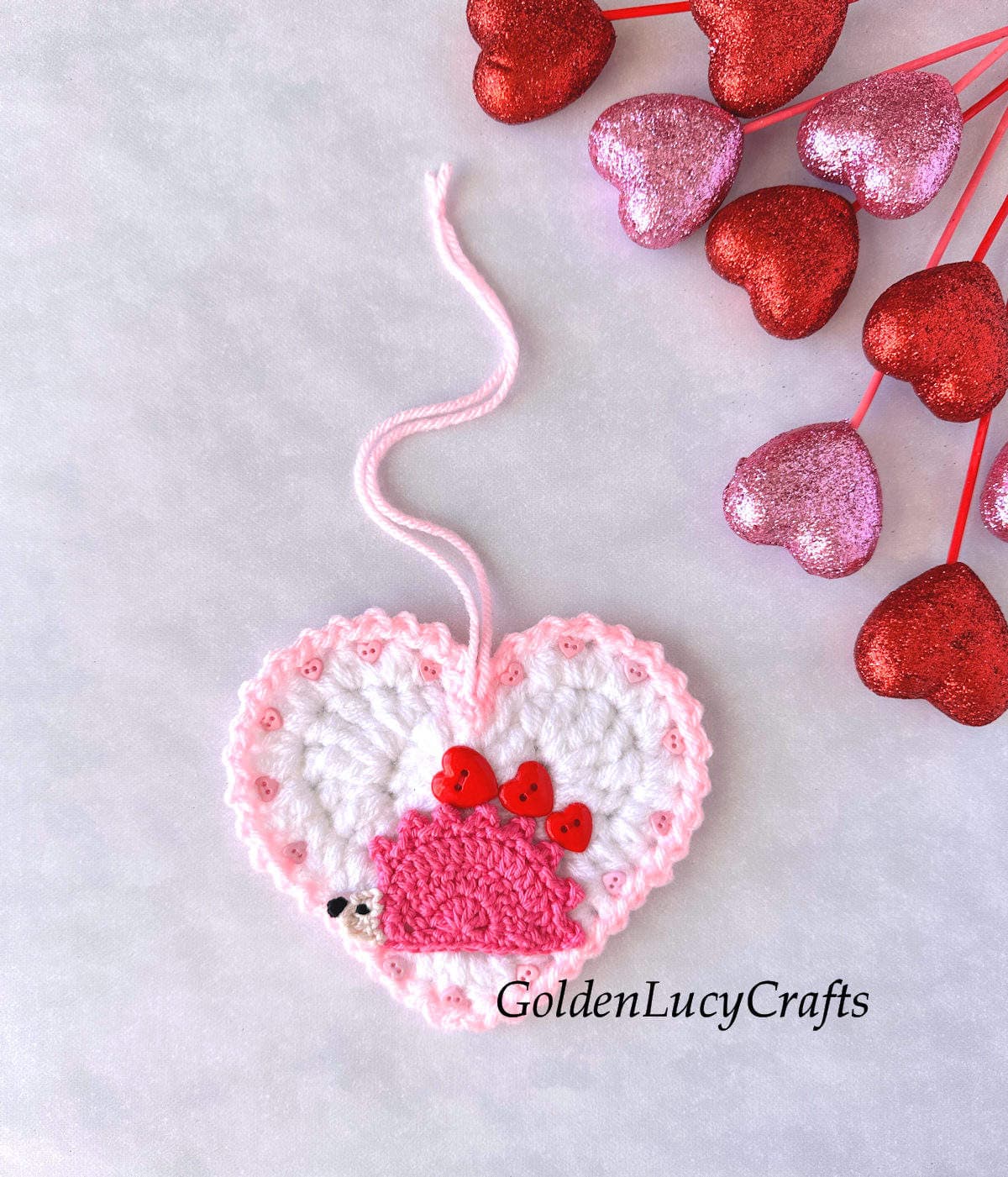 Crocheted heart-shaped ornament for Valentine's Day, featuring a pink hedgehog carrying three red hearts on its back. The ornament is made with white yarn and has a pink edge.