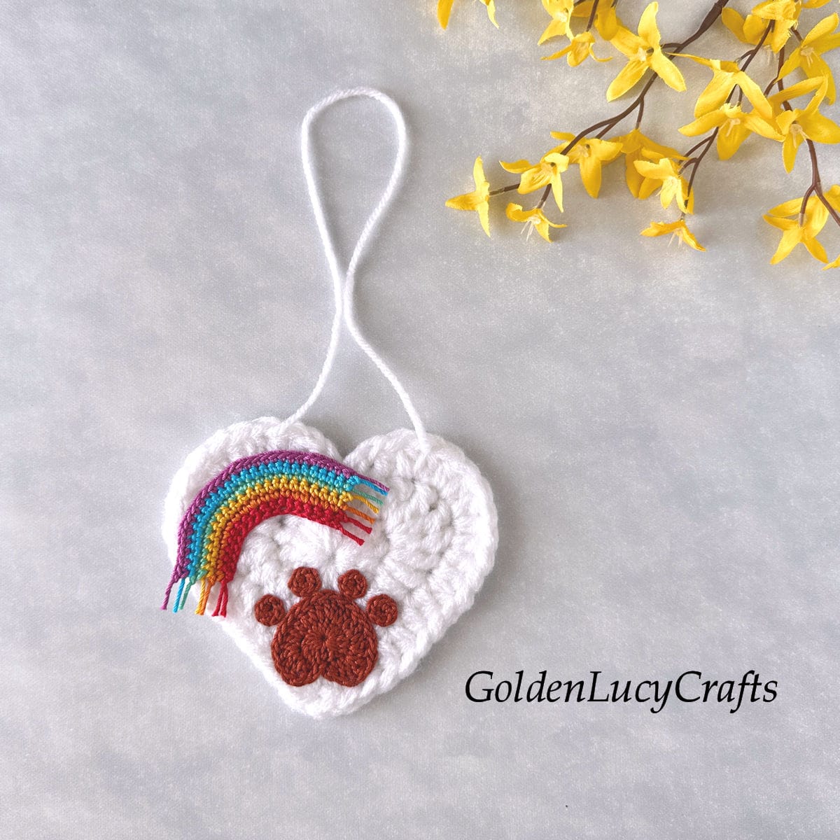 A crocheted white heart ornament with paw print and rainbow appliqués on its surface.