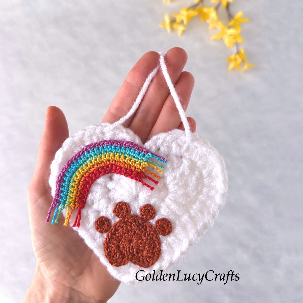 A crocheted white heart with a paw print and a rainbow on it, resting in the palm of a hand.