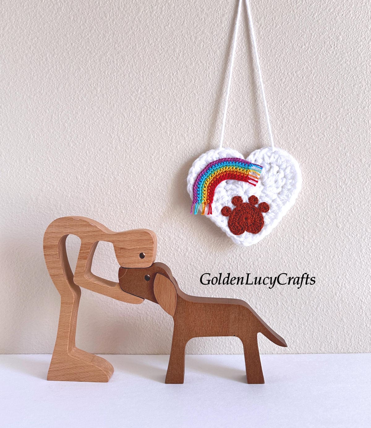 A white crocheted heart ornament hangs on the wall, featuring a paw print and a rainbow, with a wooden figurine of a man and a dog next to it.