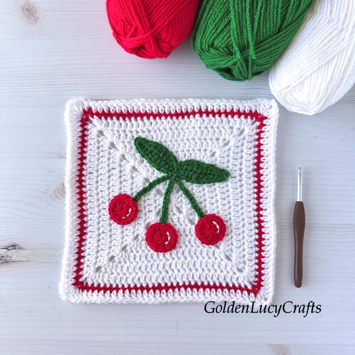 Crocheted cherry granny square, crochet hook, and three skeins of yarn in red, green, and white colors.