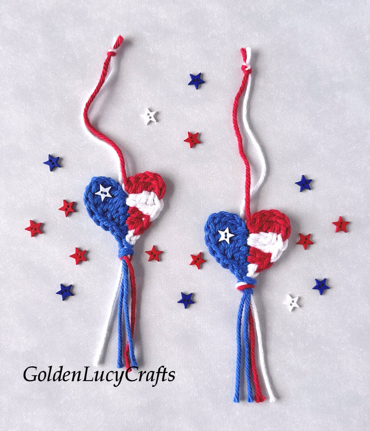Two crocheted hearts with tassels in red, white, and blue, with tiny stars lying around.