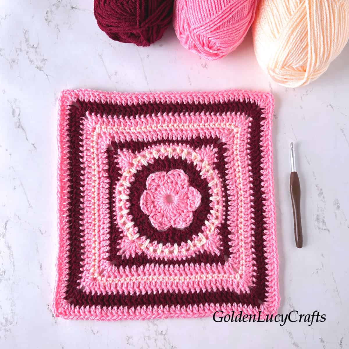 Crocheted square made in pink, dark red and cream colors, crochet hook, three skeins of yarn.