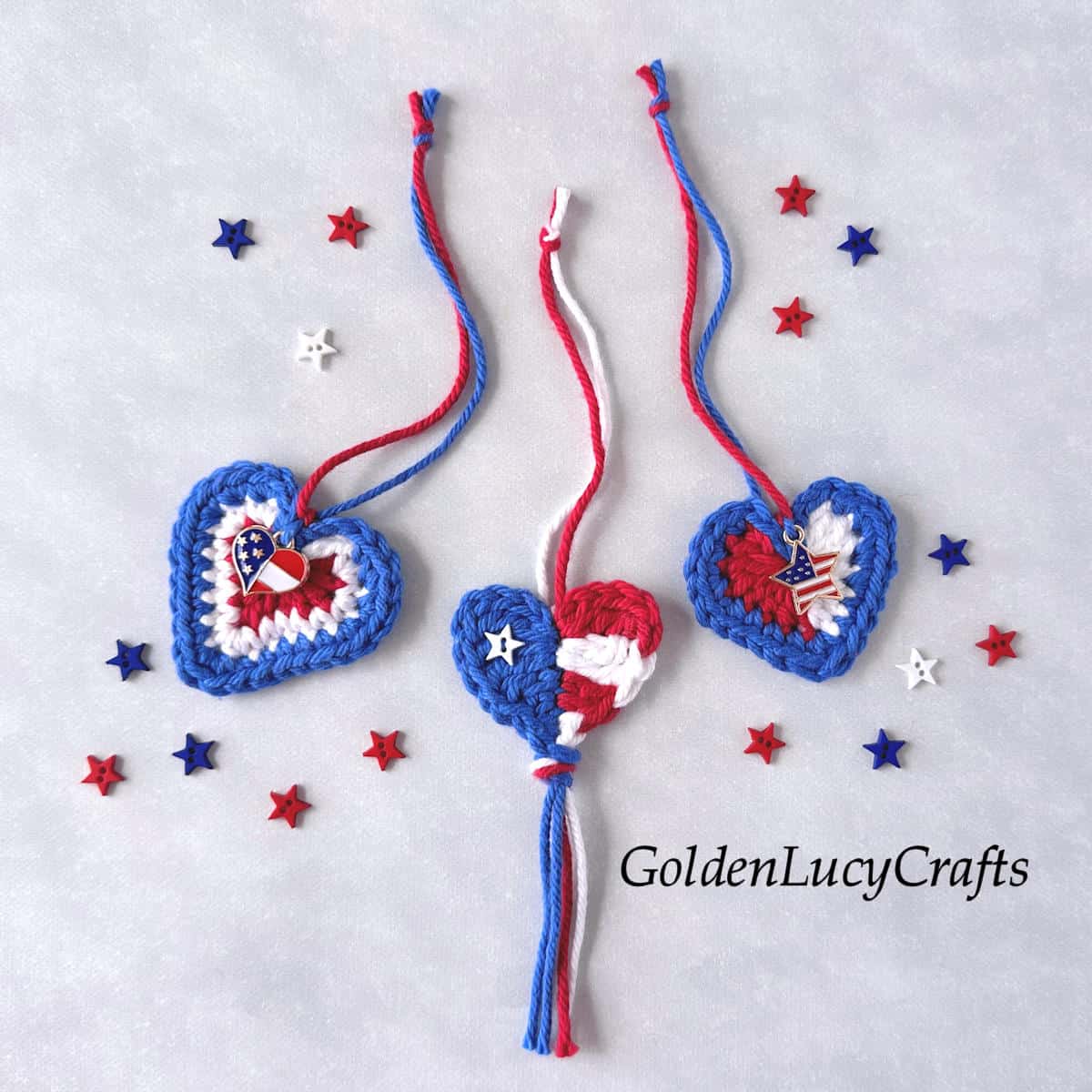 Three crocheted patriotic hearts made with red, white, and blue yarn, embellished with charms and tiny star buttons scattered around.