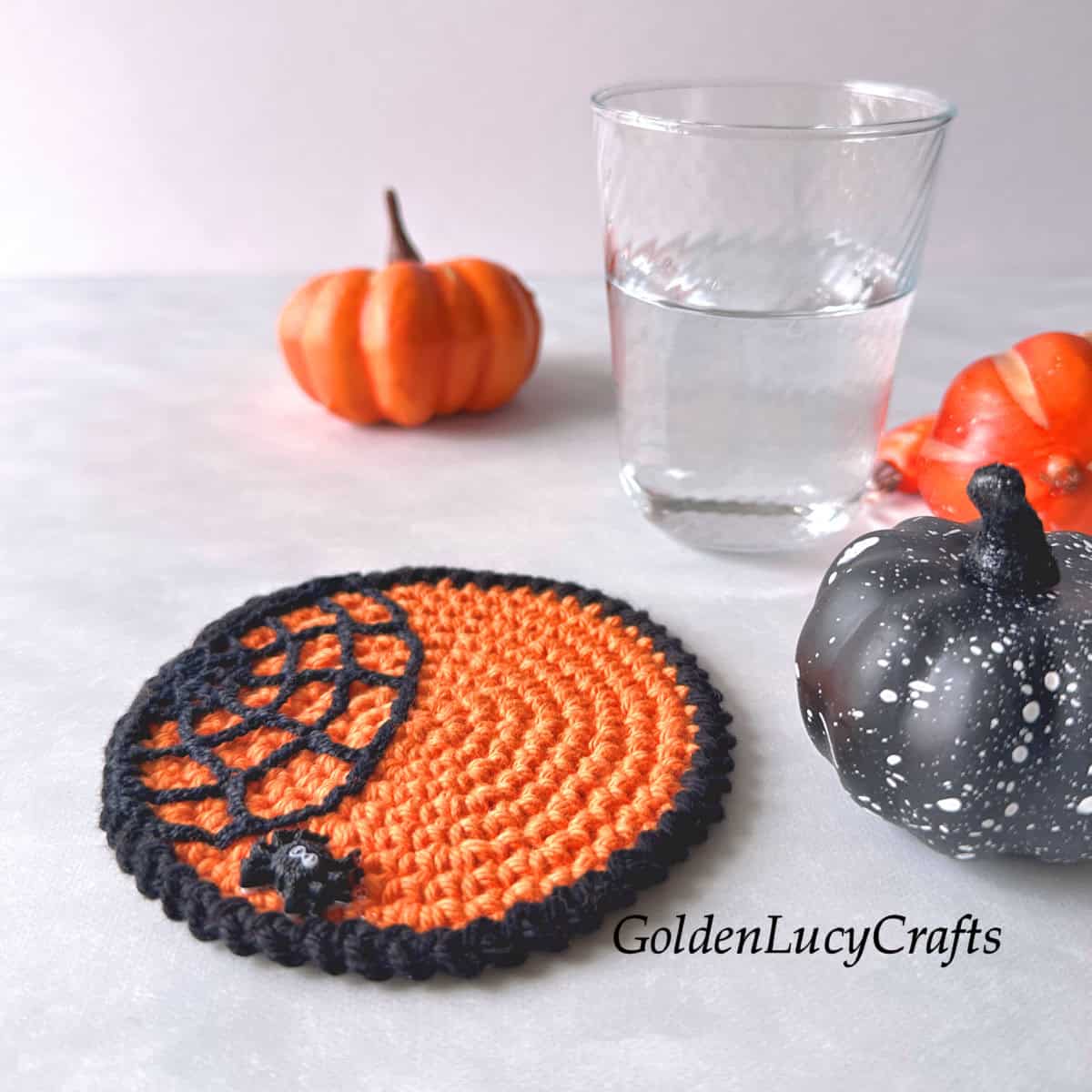 Crocheted Halloween coaster in orange, featuring a spider web with a small spider, orange and black pumpkins, and a glass of water.
