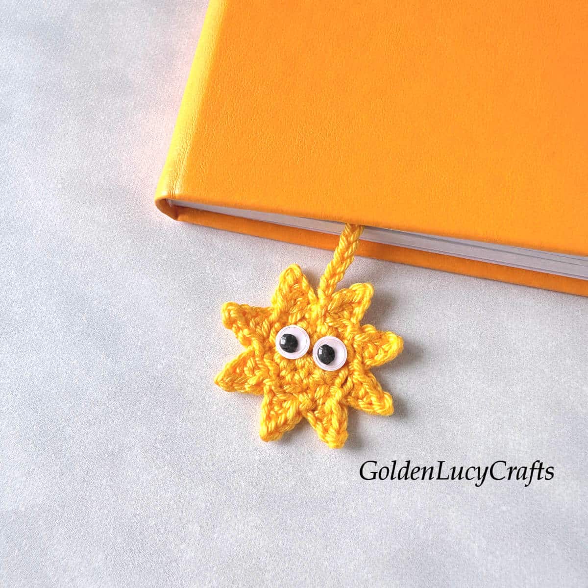 A yellow crocheted sun bookmark with googly eyes, placed into an orange book.