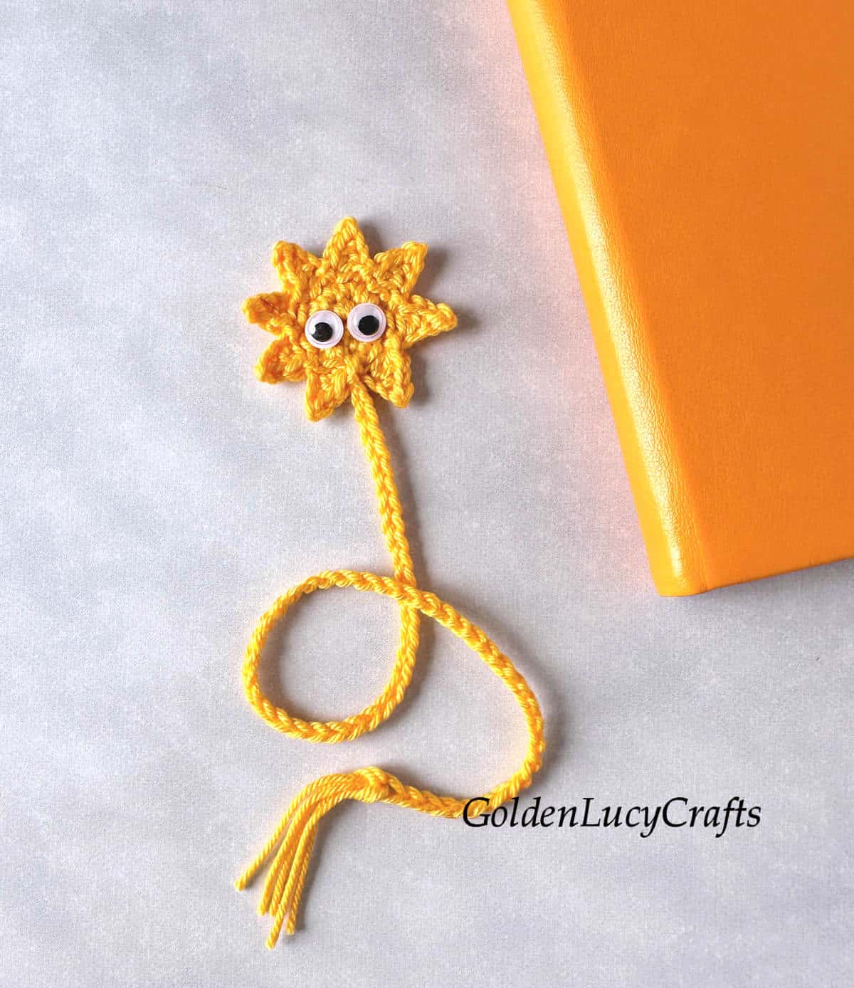 A yellow crocheted sun bookmark with googly eyes, placed next to an orange book.