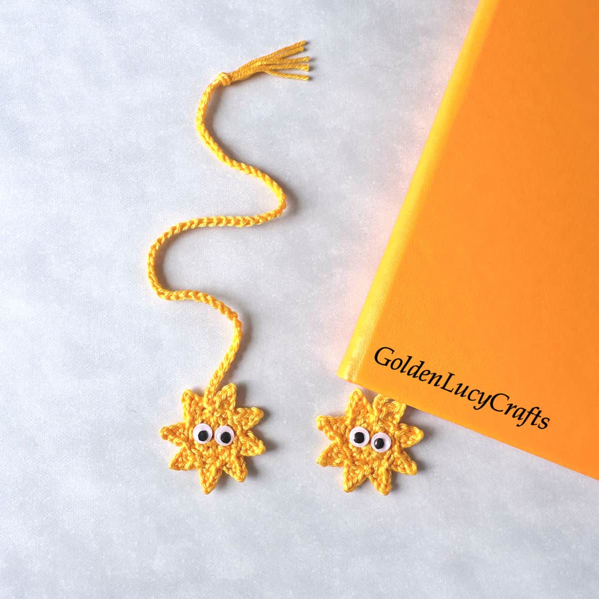 A yellow crocheted sun bookmark with googly eyes is placed into an orange book; another bookmark is next to the book.