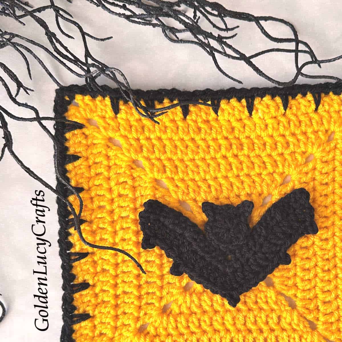 Close-up picture of a crocheted yellow square block with a black edge and a black bat at the center.