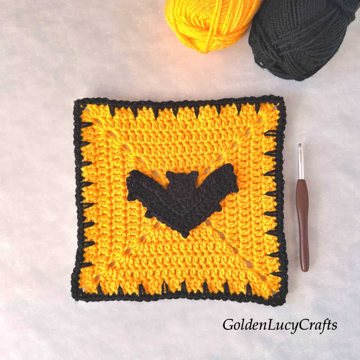 Crocheted yellow square block with a black edge and a black bat in the center, with a crochet hook and two skeins of yarn next to it.