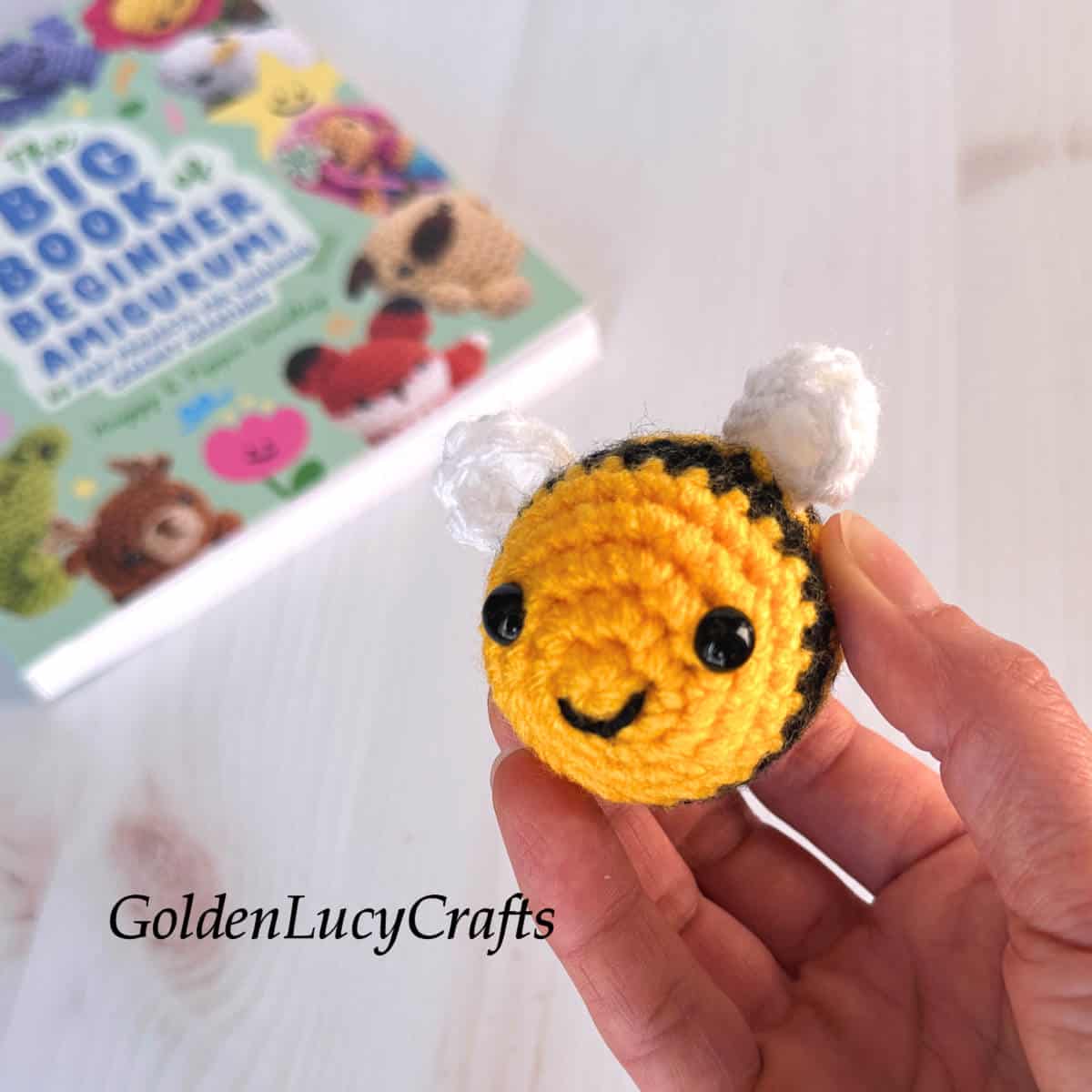 A hand is holding a small crocheted bumblebee, with a book in the background.