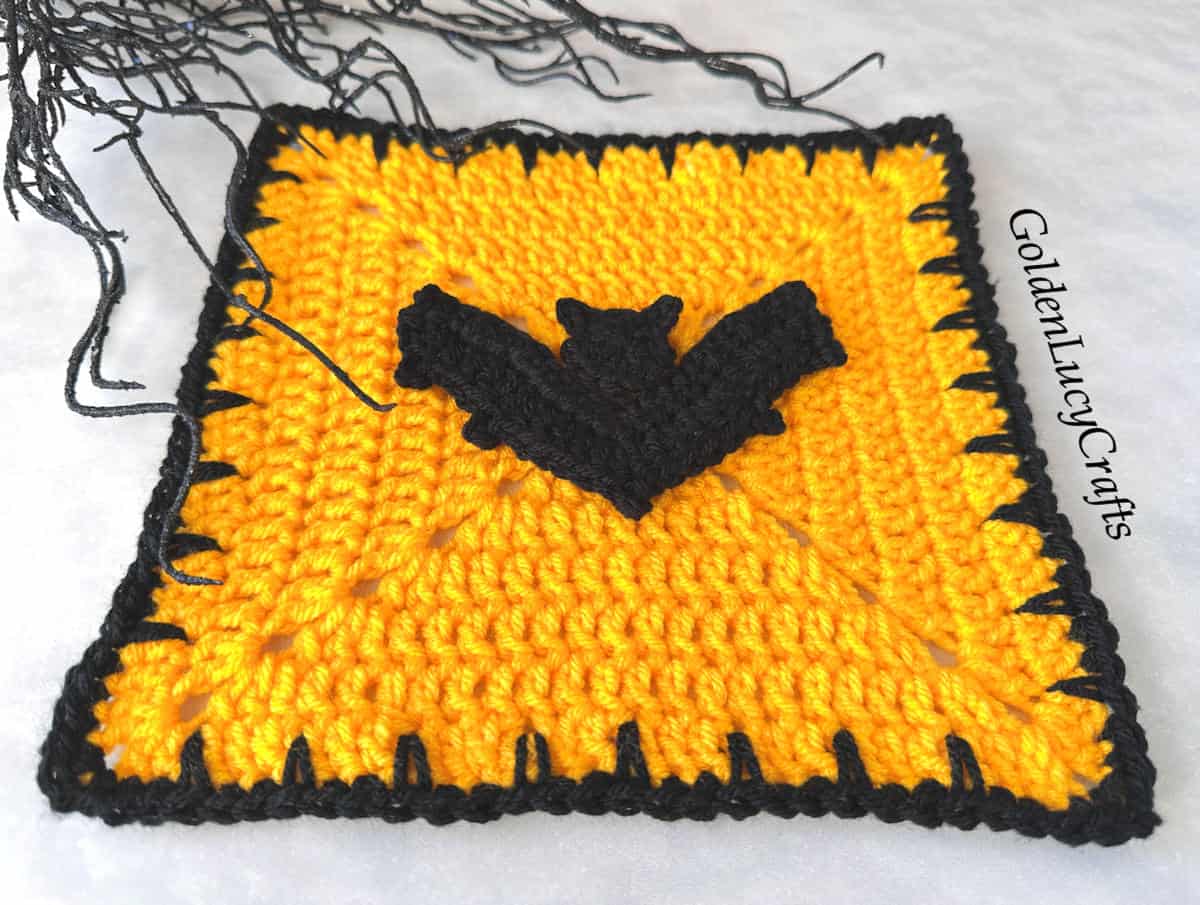 Crocheted yellow square block with a black edge, a black bat in the center, and black branches in the background.